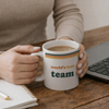 A person holding a cup that says 'world's best team' while sitting at a wooden desk with a laptop and notepad.