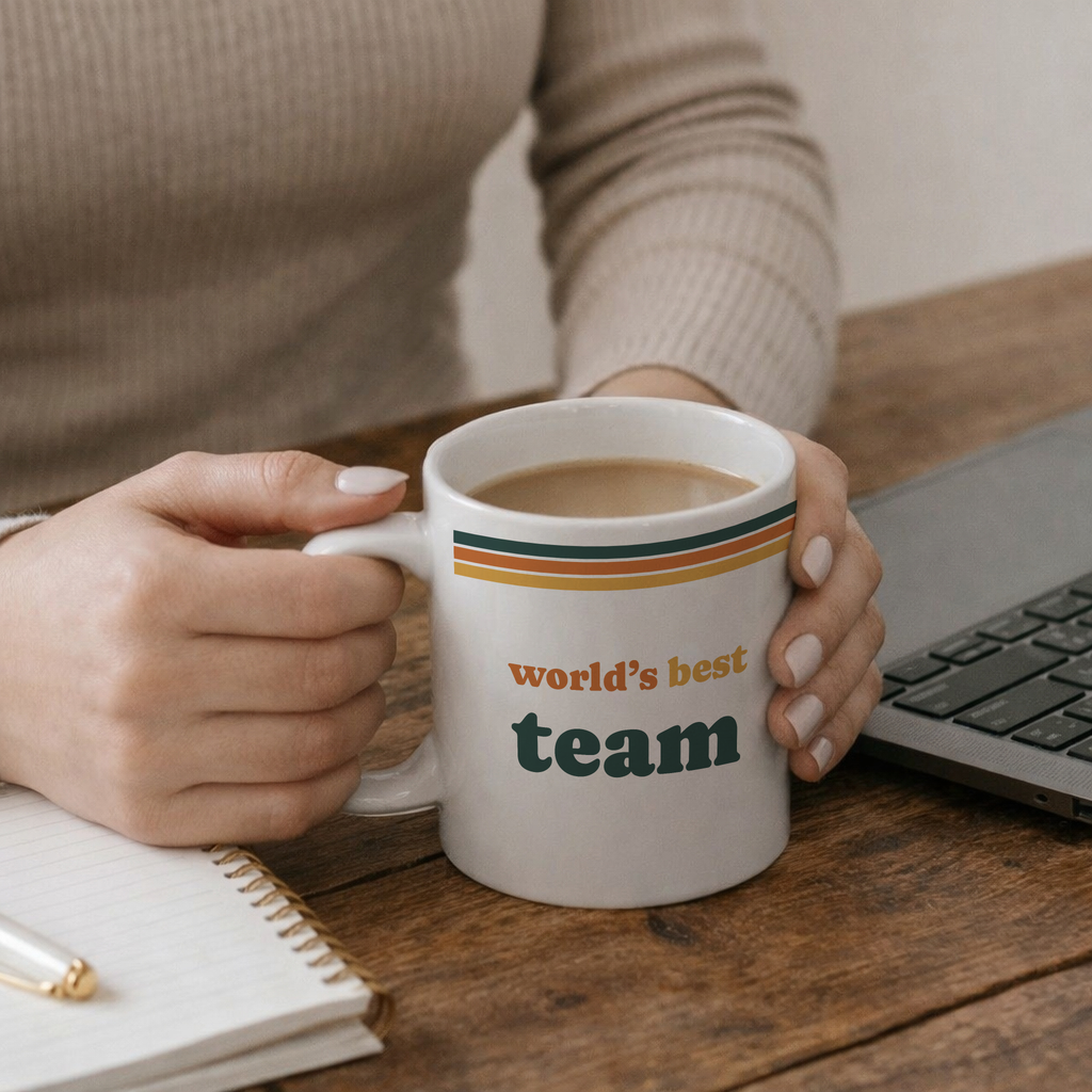 A person holding a cup that says 'world's best team' while sitting at a wooden desk with a laptop and notepad.